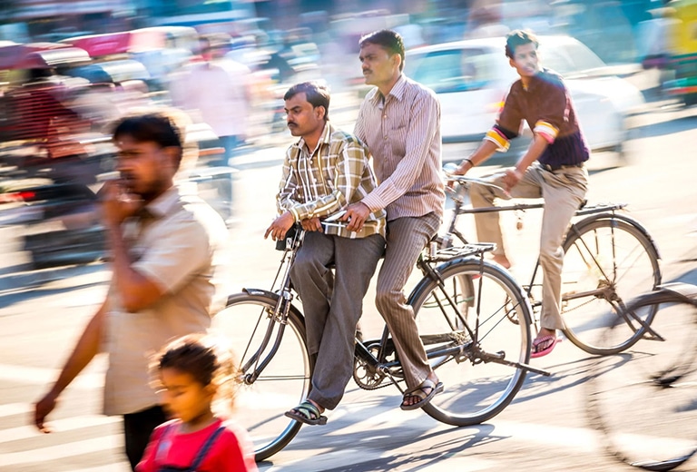 3 hombres de etnia hindú que van montados en dos bicicletas, mientras otros caminan por una calle